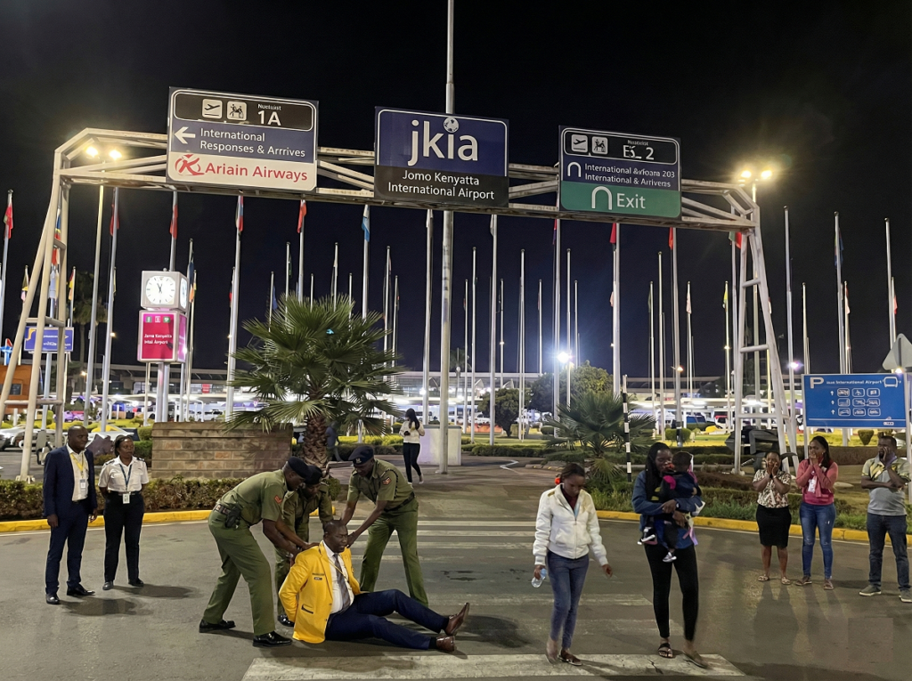 Three uniformed officers arrest a man wearing a bright yellow blazer on the tarmac outside the Jomo Kenyatta International Airport (JKIA) terminal entrance at night, while several bystanders look on in shock.