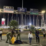 Three uniformed officers arrest a man wearing a bright yellow blazer on the tarmac outside the Jomo Kenyatta International Airport (JKIA) terminal entrance at night, while several bystanders look on in shock.